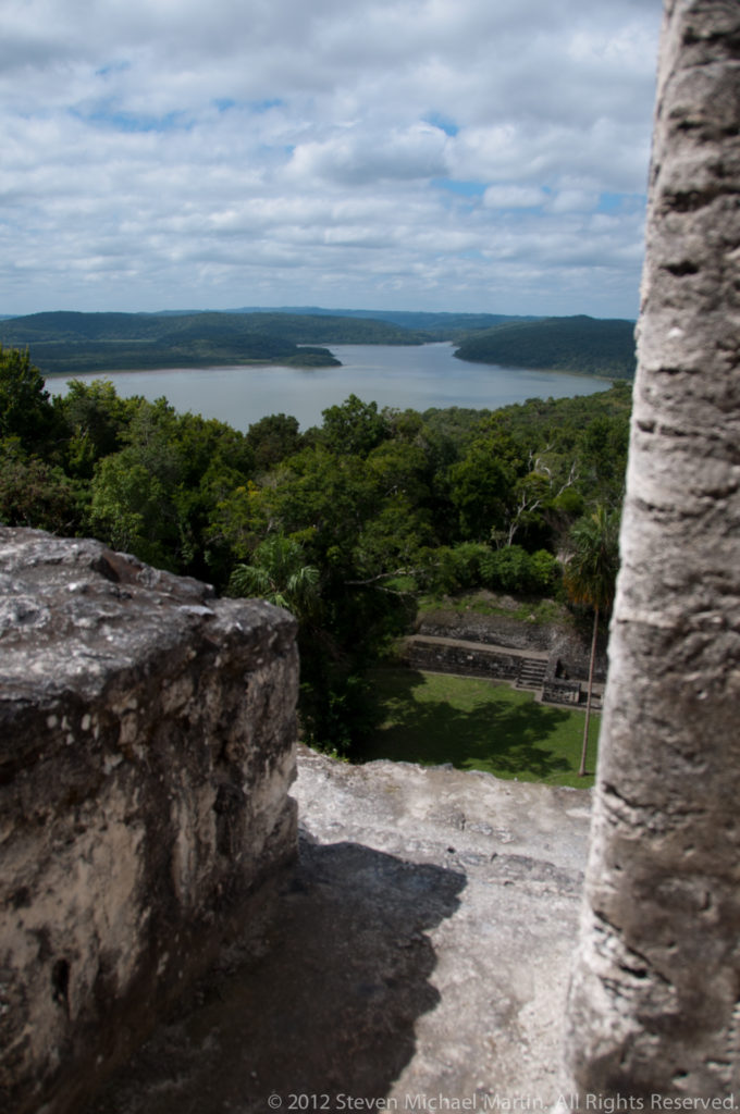 Elevated view from Maya stonework across tropical forest to Lake Yaxha beneath a cloudy sky.