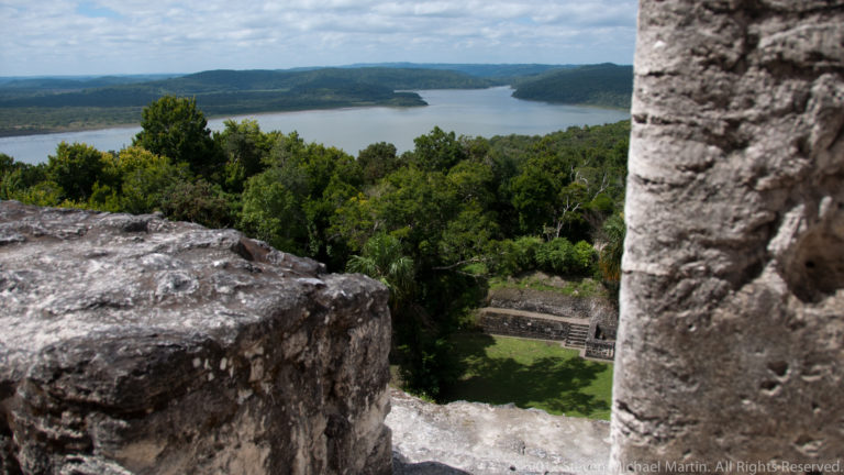 High vantage over stone ruins and a green forest canopy toward a broad lake, with a lower plaza visible below.
