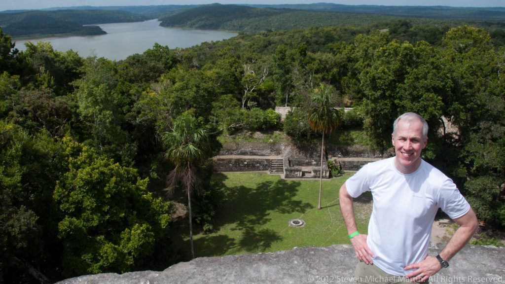 Man in a white shirt standing on a high stone structure above Yaxha, with forest, plaza, and lake behind him.