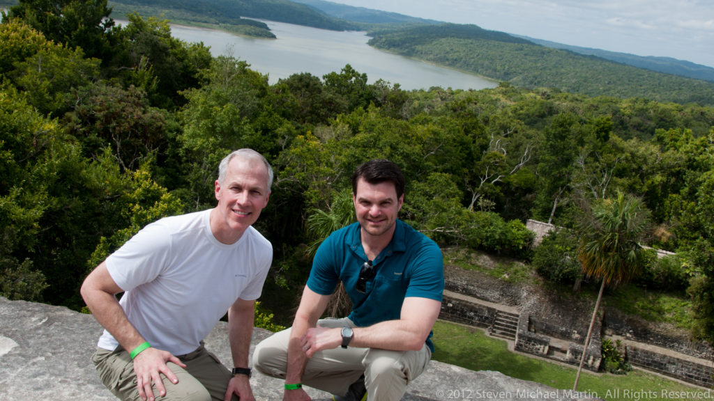 Two men seated on an elevated Maya structure above a forested plaza and lake at Yaxha.