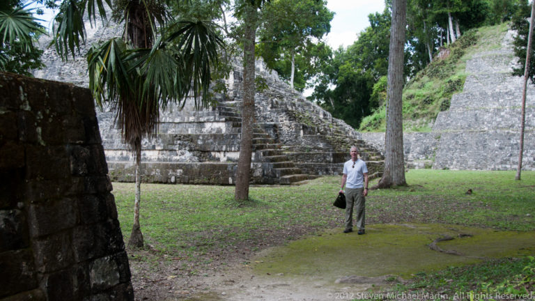 Man standing in a grassy archaeological court with stepped stone structures and tropical trees around him.