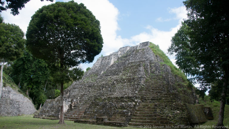 Large stepped stone pyramid with a central stairway rising between mature trees at Yaxha.