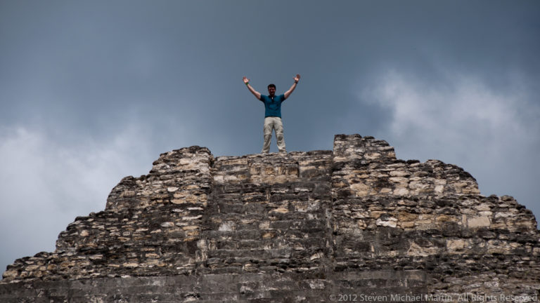 Man with raised arms standing atop a weathered stone Maya structure beneath a dark cloudy sky.
