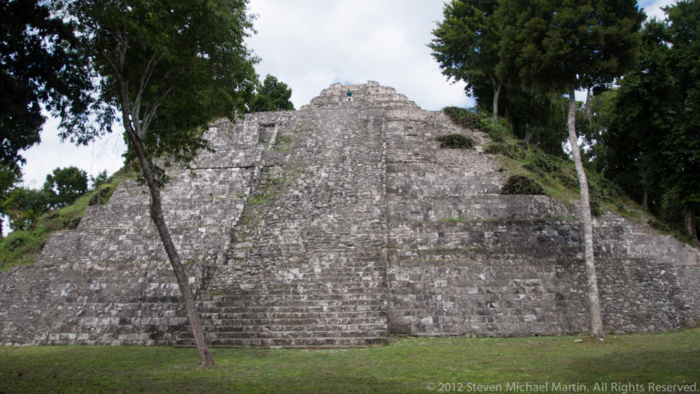 Tall stepped stone pyramid with a broad central stairway rising from grass between trees at Yaxha.