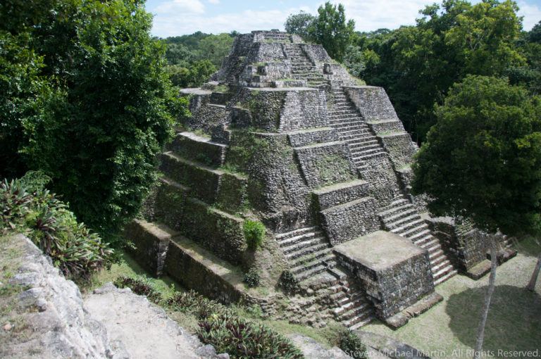 Oblique elevated view of a stepped Maya pyramid with several stairways and terraces surrounded by dense green forest.