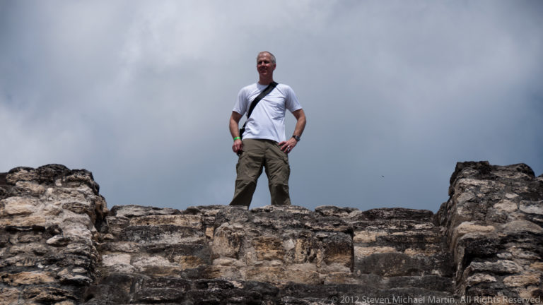 Visitor standing above weathered Maya stone walls at Yaxha beneath a cloudy sky.