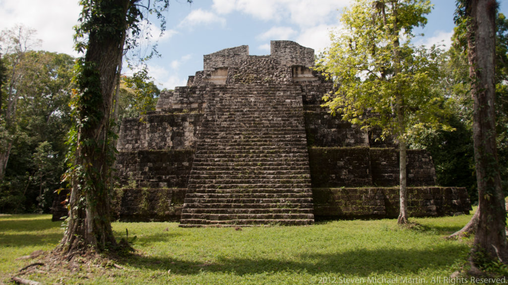 Stepped Maya temple-pyramid with a broad central staircase rising from a grassy plaza at Yaxha.