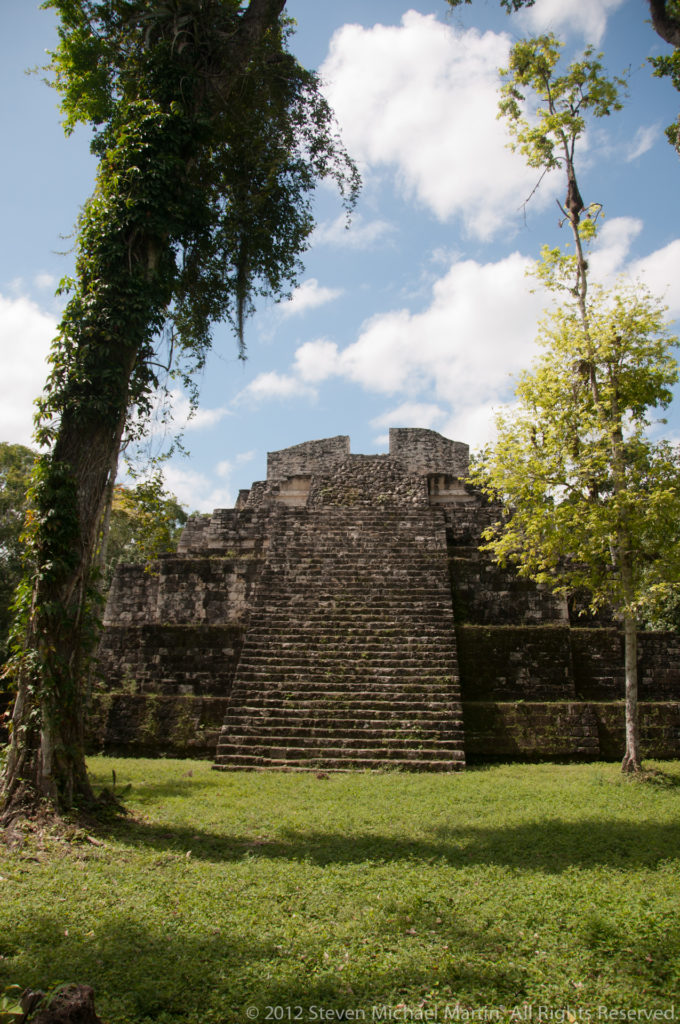 Wide view of a stepped Maya temple-pyramid across a grassy plaza with trees at Yaxha.