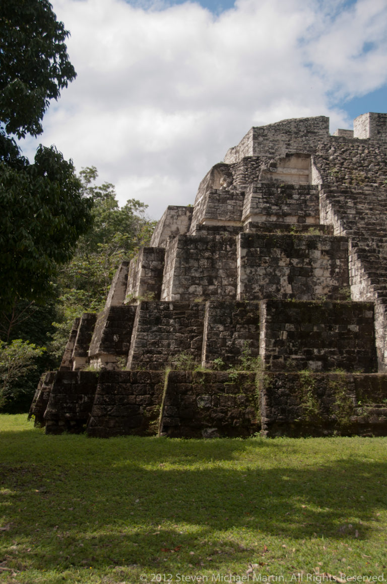 Oblique view of a terraced Maya temple-pyramid with weathered stone steps beside a grassy lawn.