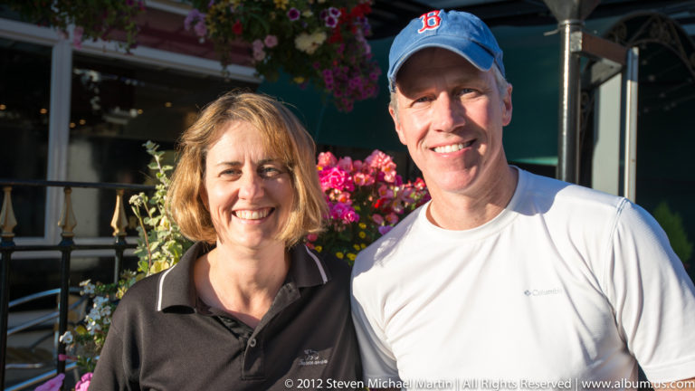 Two smiling visitors pose beneath a canopy, with bright flowers and leafy planting behind them.