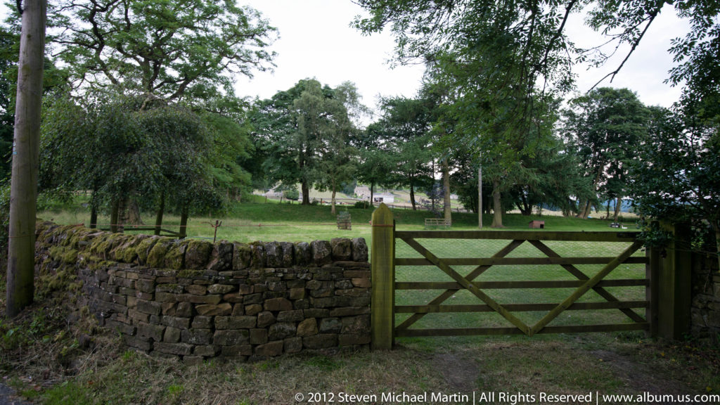 A braced wooden gate closes a gap in a dry-stone wall, with a tree-dotted pasture beyond.