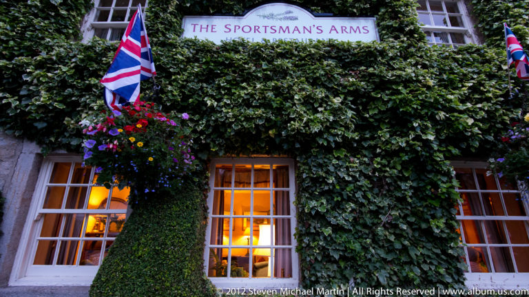 The Sportsman’s Arms sign rises above two warmly lit windows framed by ivy and hanging flowers.