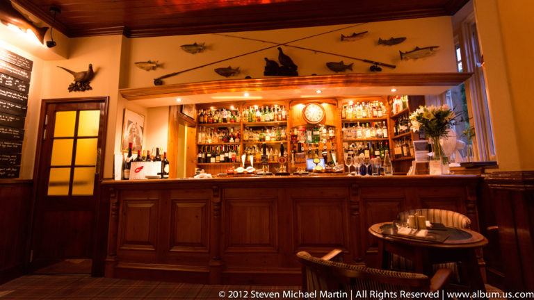 A wood-paneled pub bar displays bottles beneath a clock and mounted fish, with a small table in front.