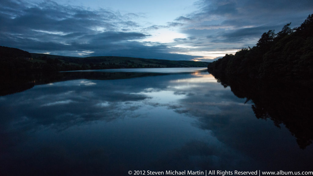 A calm lake mirrors dark wooded shores and a blue dusk sky with a pale opening at the horizon.