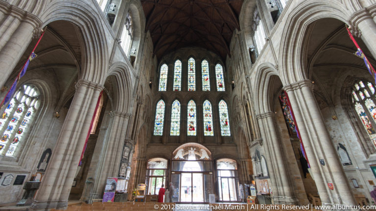 Pointed arches and stone columns frame a church nave ending at tall stained-glass windows and a distant doorway.