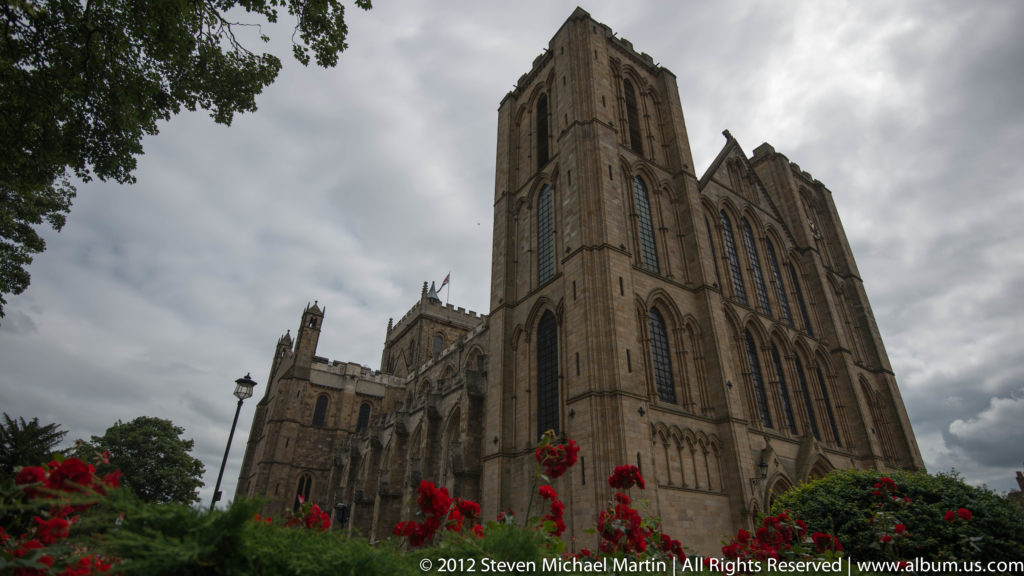 A square stone church tower with narrow windows rises behind red flowers under a cloudy sky.