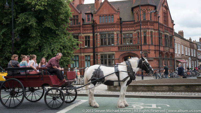 A white horse pulls a red passenger carriage past pedestrians and a red-brick town building.
