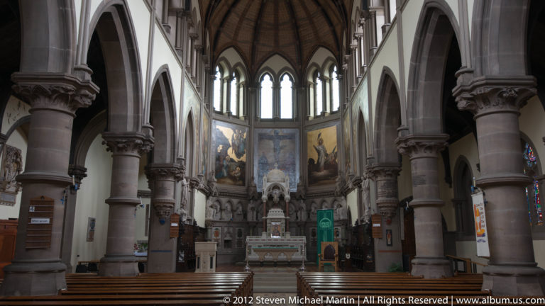 Rows of chairs lead through a stone church nave toward a distant altar beneath pointed arches and stained glass.