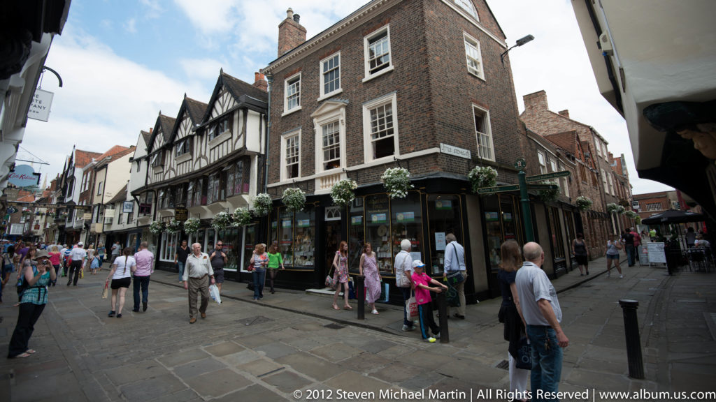 Pedestrians walk beside black-and-white timber-framed shopfronts and brick commercial buildings on a paved street.