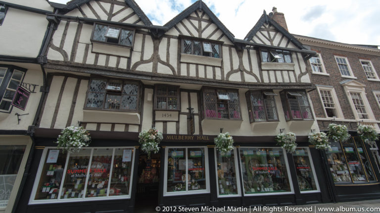 Dark beams outline projecting upper stories above modern shop windows and flower baskets.
