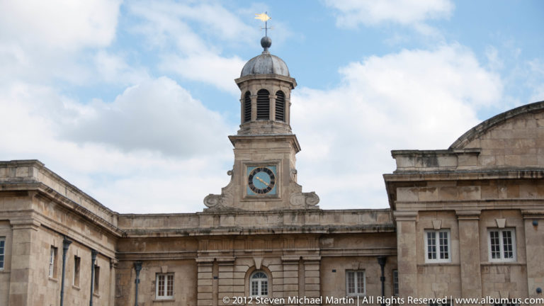 A round clock sits in a small domed cupola above a pale stone façade and neighboring rooftops.