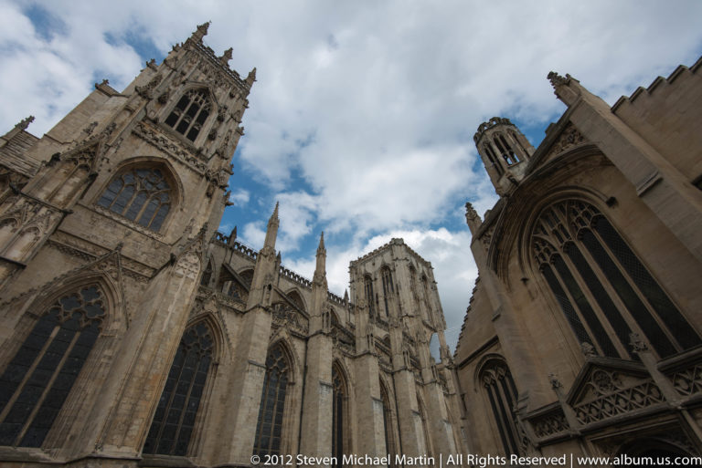 Pale stone Gothic buildings with pointed windows and pinnacles rise around a narrow strip of cloudy sky.