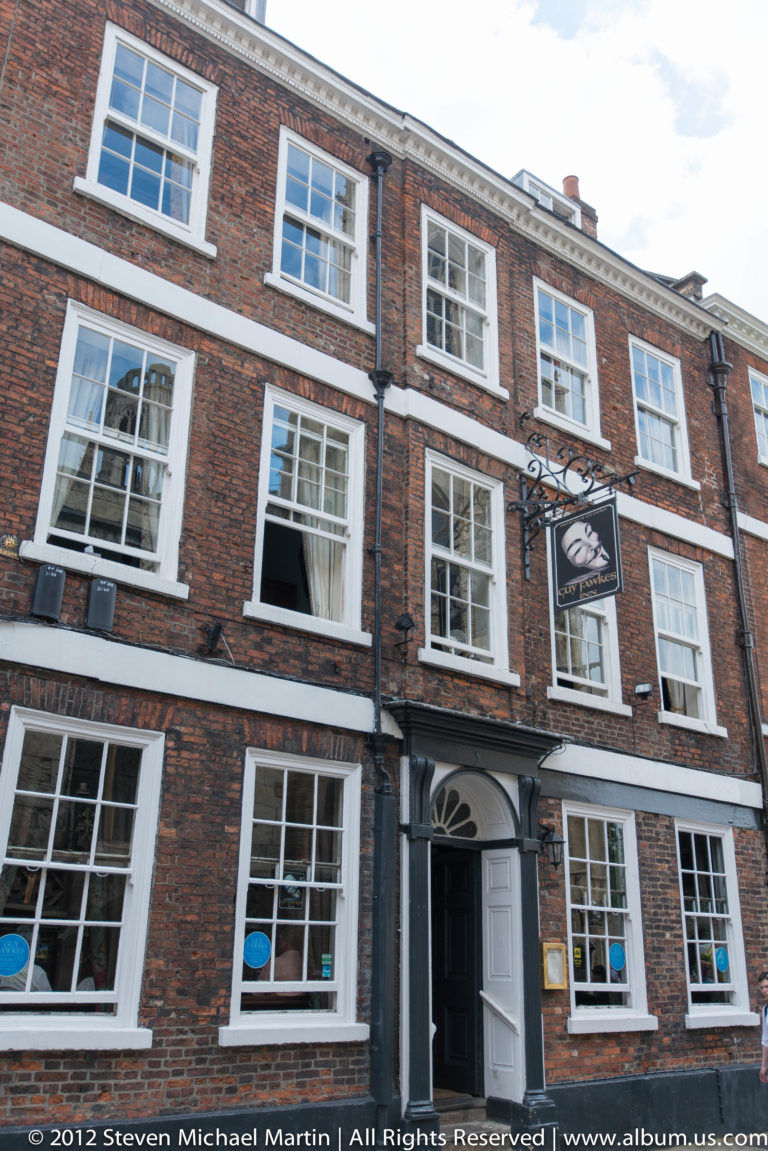 A red-brick pub has white-trimmed upper windows and a hanging sign above its street-level entrance.