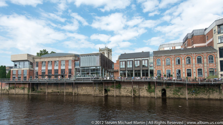 Brick and modern commercial buildings line a broad river above a raised walkway and masonry supports.