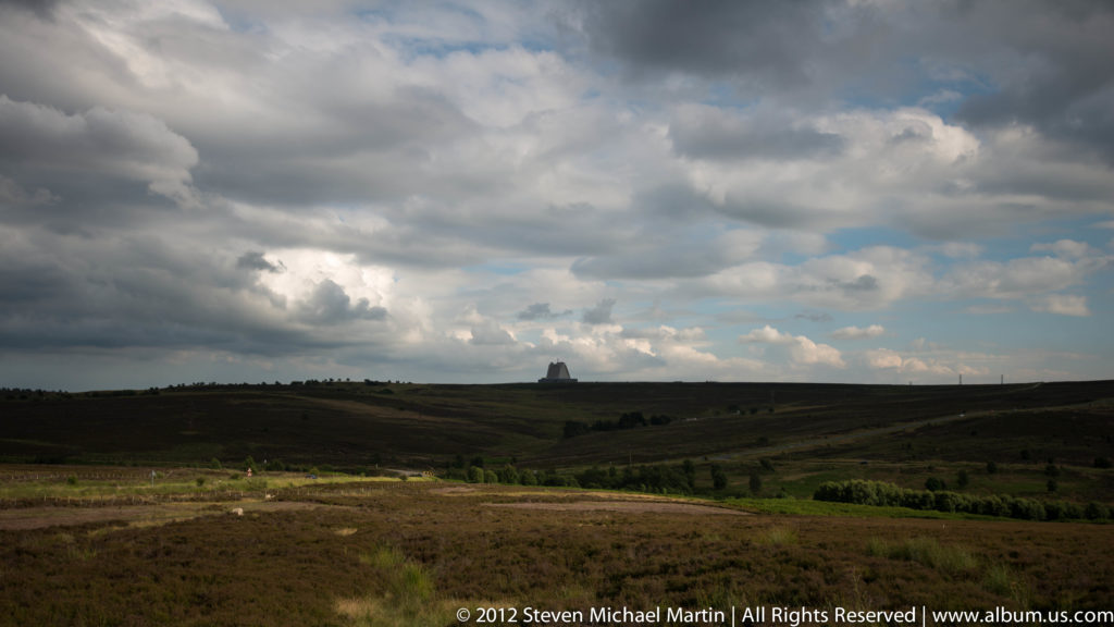 Open fields extend toward a distant tower beneath layered clouds and a bright patch of sky.
