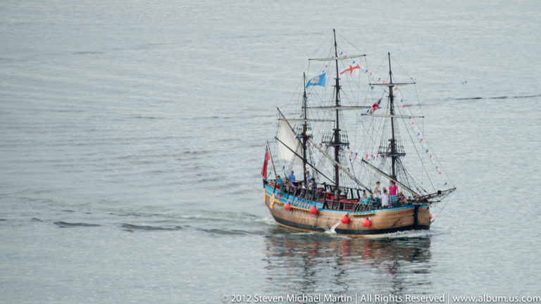 A dark-hulled tall ship with several masts and dense rigging crosses calm water beneath a gray sky.