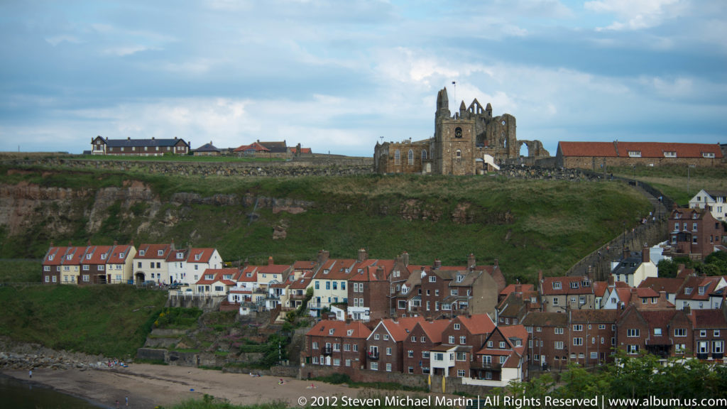A stone church tower rises above clustered red-roofed houses along a coastal harbor.