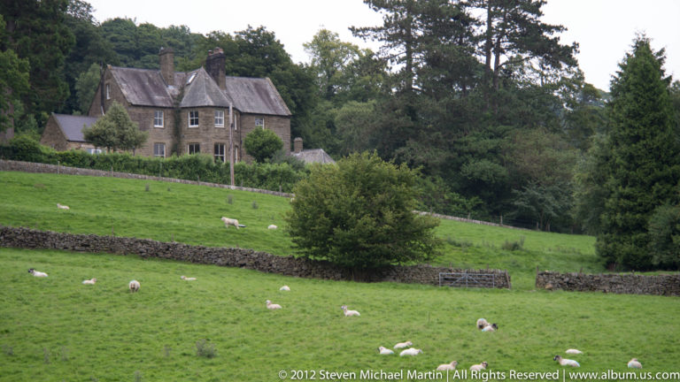 Sheep graze across a walled green slope below a large stone house partly screened by trees.