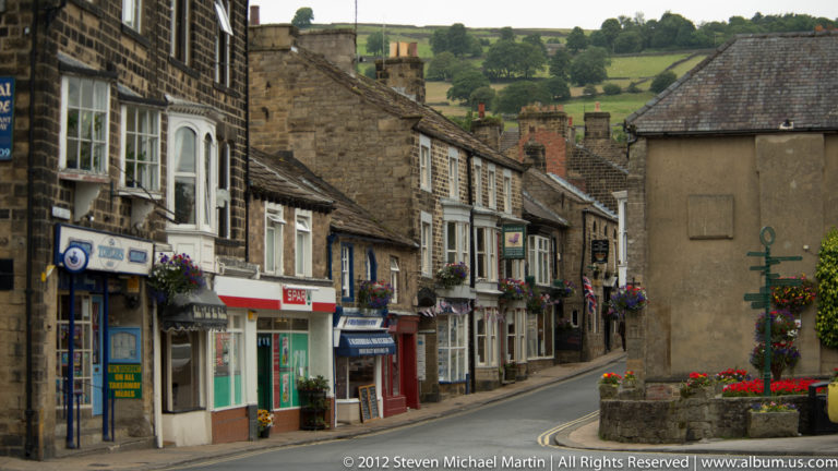 Stone shopfronts line a curving town street that leads toward green hills.