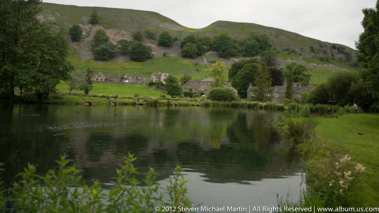 Stone houses stand across a calm lake below rounded green hills and scattered trees.