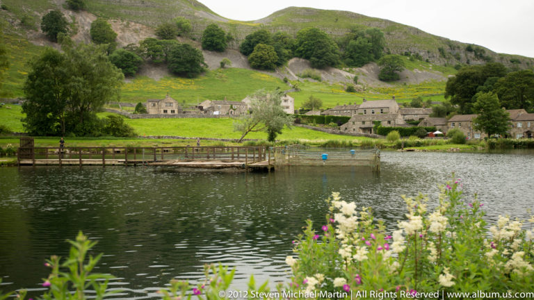 A wooden landing reaches across a lake toward stone houses and green hills, with flowers along the near bank.