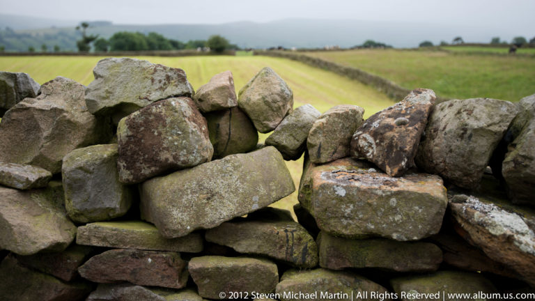 Rough irregular stones form a close-up dry-stone wall with a blurred green field beyond.