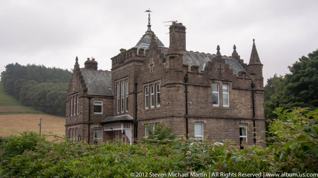 A turreted stone country house with steep roofs and chimneys stands beyond a lawn and flower border.
