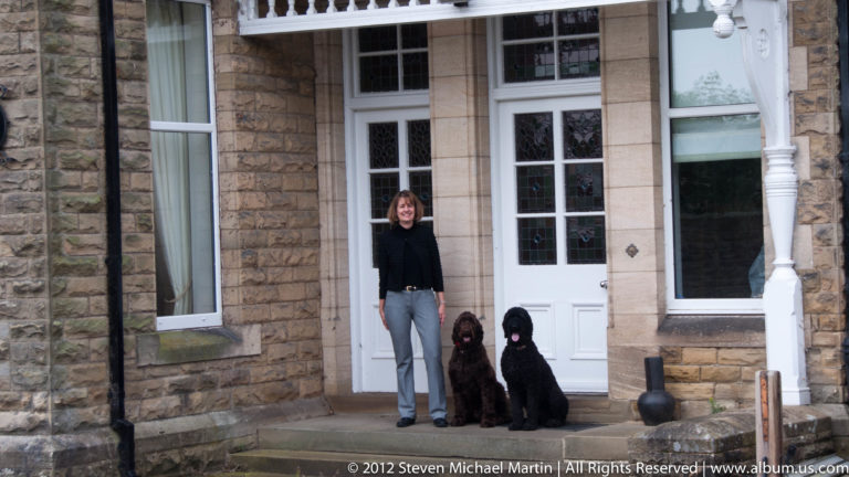 A woman stands beside a brown dog and a black dog at a white doorway in a stone building.
