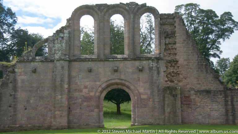 Three tall window openings rise above an arched doorway in a ruined abbey wall.