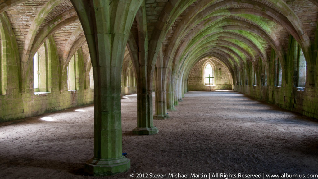 A vaulted stone corridor at Fountains Abbey recedes through repeated arches toward a window.