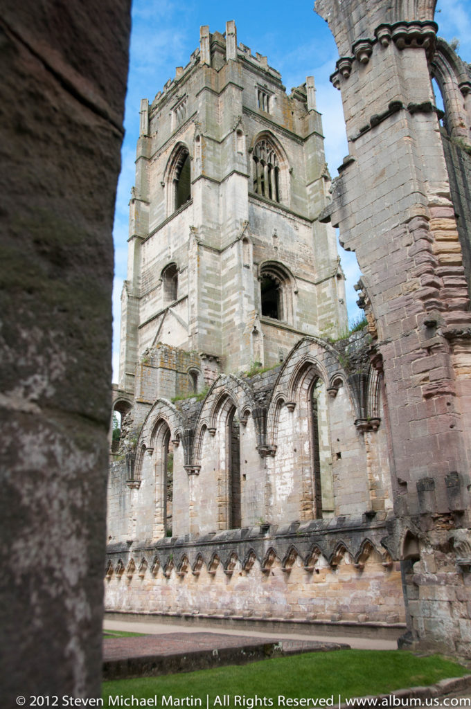 A high stone abbey façade with arched windows is seen through a dark foreground arch.