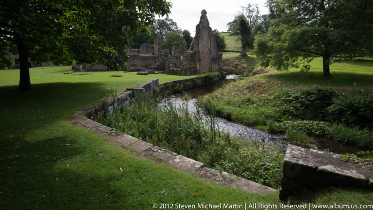 A water channel and low stone wall lead toward abbey ruins in a green garden.