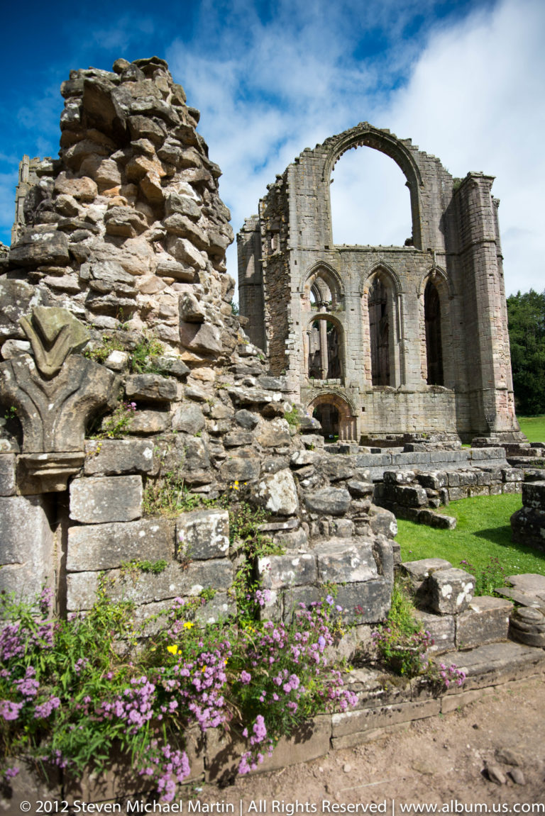 Purple flowers and low foundations lead toward a roofless abbey gable.