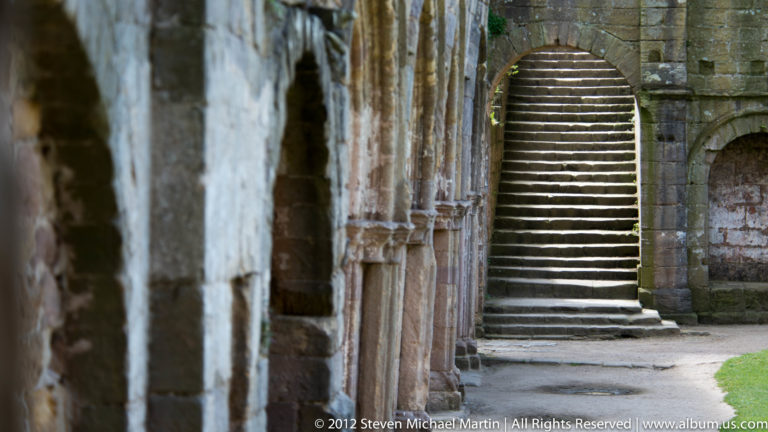 Stone steps rise through a narrow arched doorway in a thick abbey wall.