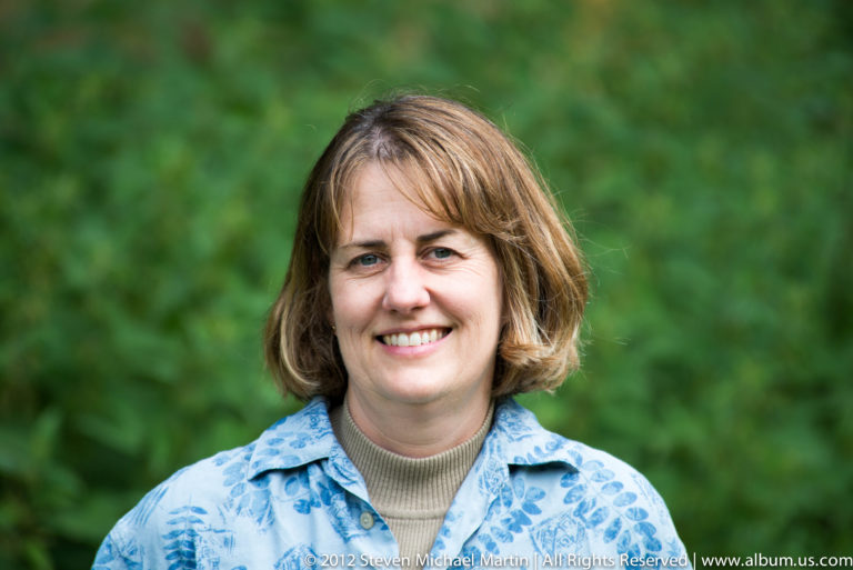 A woman in sunglasses and a blue patterned shirt poses against green foliage.