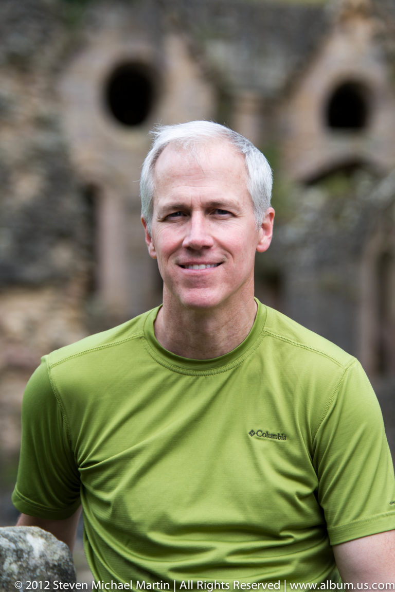 A white-haired man in a lime-green shirt smiles at the camera, with blurred abbey stonework behind him.