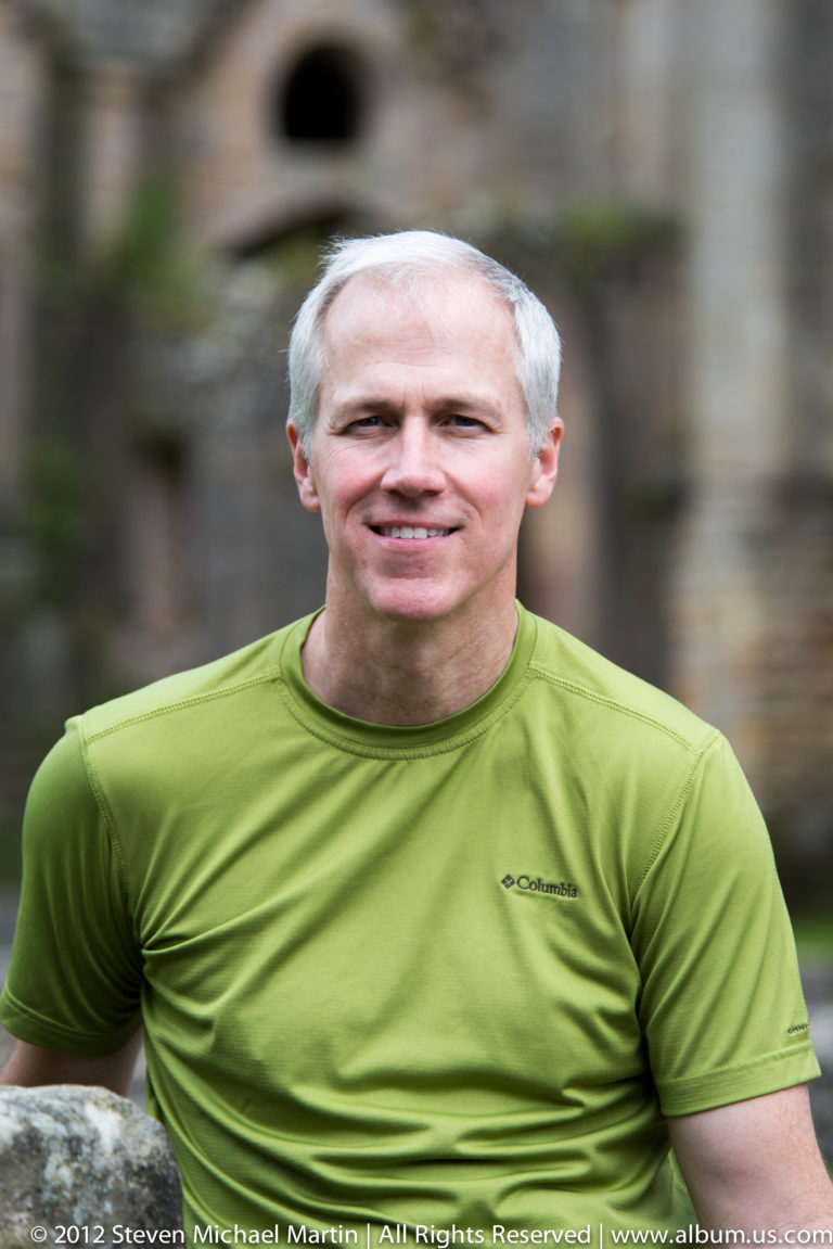 A smiling white-haired visitor in a lime-green shirt is shown chest-up against dark, blurred stone and greenery.