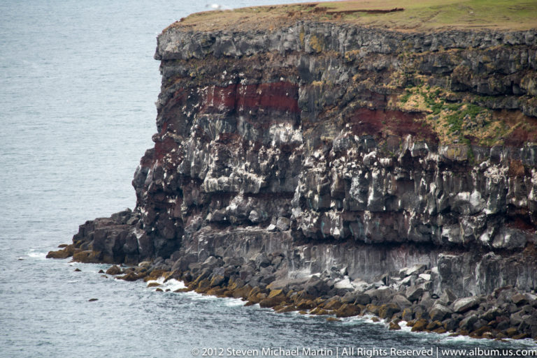 Columnar basalt cliff with a grassy rim rises above angular shoreline rocks and the North Atlantic.