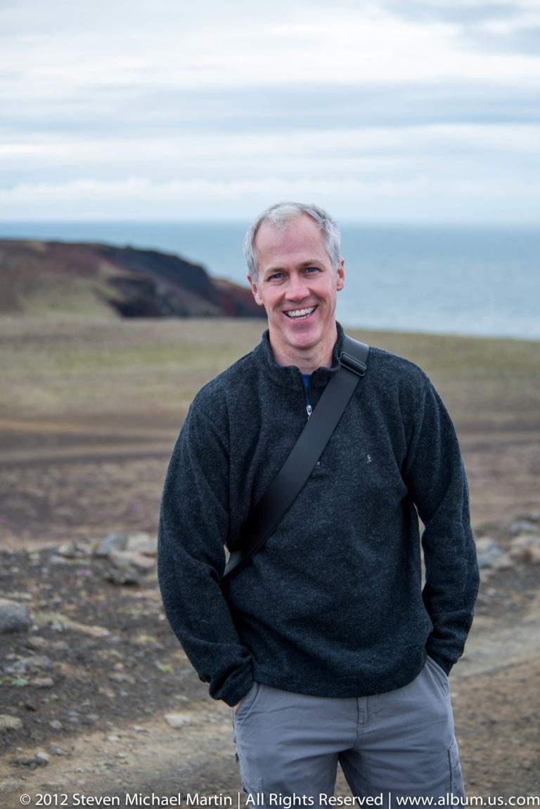 A smiling traveler in a dark sweater stands before a low coastal headland under a pale sky.