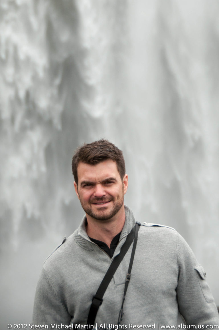 A smiling man in a dark jacket stands against a pale sky above a narrow strip of sea.